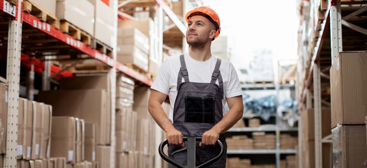Homem em um ambiente de trabalho de armazenamento industrial. Ele está usando uniforme e capacete, o que sugere que ele está em um local onde a segurança é importante. O homem está operando um carrinho hidráulico, que é um equipamento usado para mover objetos pesados. Ele está em um armazém, que é um grande espaço cheio de caixas. As caixas sugerem que o local é usado para armazenar e transportar mercadorias. A cena geral sugere um ambiente de trabalho ativo e movimentado, onde o homem está desempenhando um papel importante na movimentação de materiais.